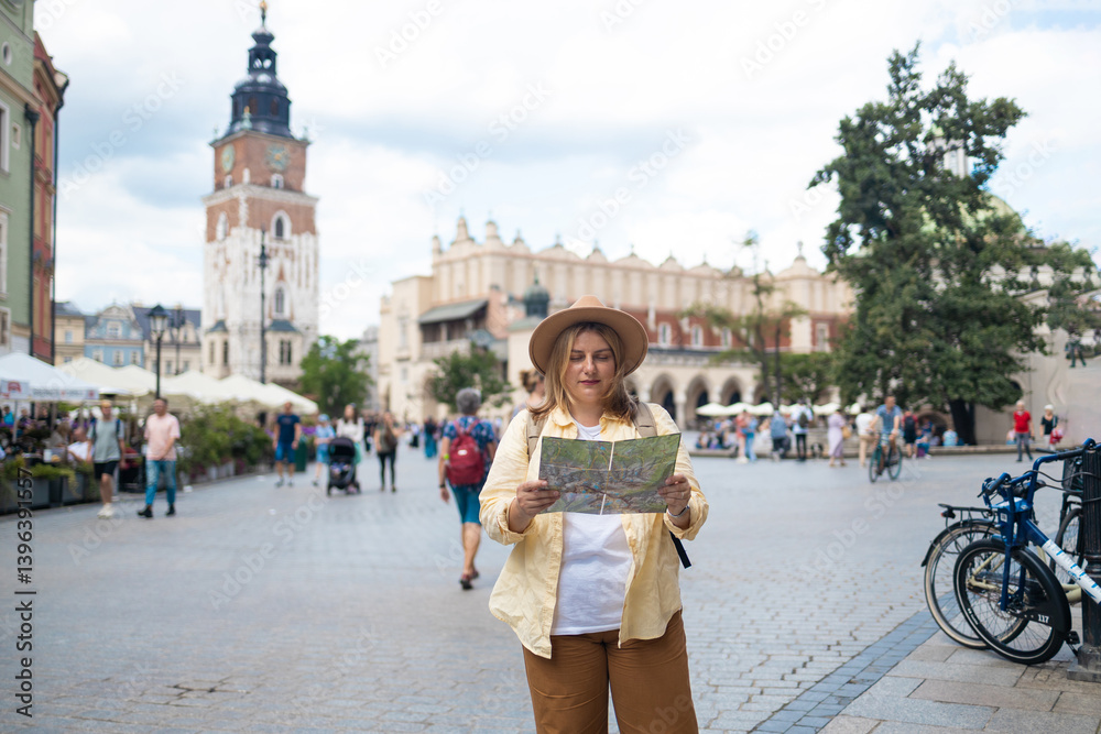 A woman wearing a yellow jacket and hat stands in a lively city square, holding a map as she tries to navigate the area. She appears to be a tourist, with the city architecture and cyclists in the