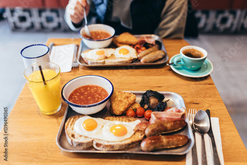 POV shot of a full English breakfast on metal trays: fried eggs, sausages, baked beans, hash browns, bacon, mushrooms, tomatoes, juice, and coffee. Soft focus