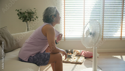 Asia adult people middle age old woman sit at sofa home feel hot flash fever use fan waving hand blowing wind face elder health care summer heat aging issue. Sad pain sweat tired stroke suffer stress.