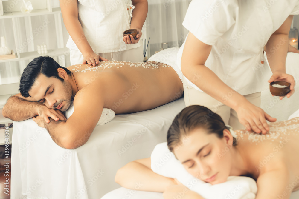 Blissful couple customer having exfoliation treatment in luxury spa salon with warmth candle light ambient. Salt scrub beauty treatment in health spa body scrub. Quiescent