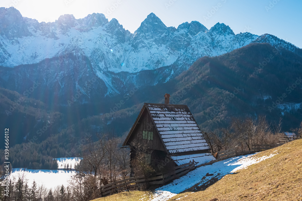 Traditional wooden cabin with snow-dusted roof rests on sunlit grassy slope, overlooking winter valley beneath majestic, snow-covered peaks of Martuljek mountain range in remote Slovenian Julian Alps