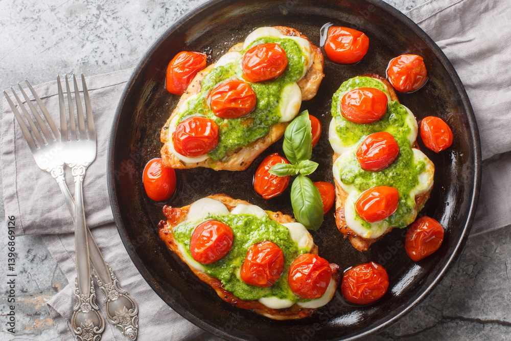 Chicken margherita is made with juicy pan-seared chicken, topped with basil pesto, mozzarella, roasted cherry tomatoes close-up on pan on table. Horizontal top view from above
