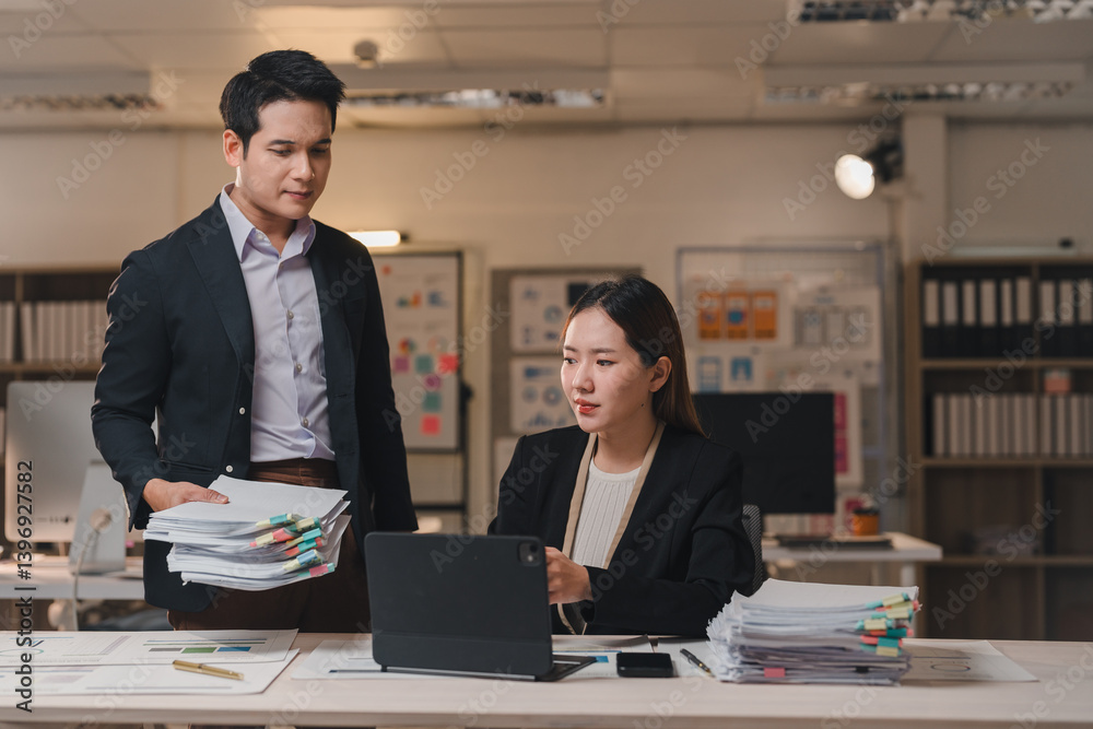 Two asian business professionals collaborating late in the office, utilizing a tablet while reviewing a stack of documents, focused on meeting project deadlines and achieving success Two asian business professionals collaborating late in the office, utilizing a tablet while reviewing a stack of documents, focused on meeting project deadlines and achieving success