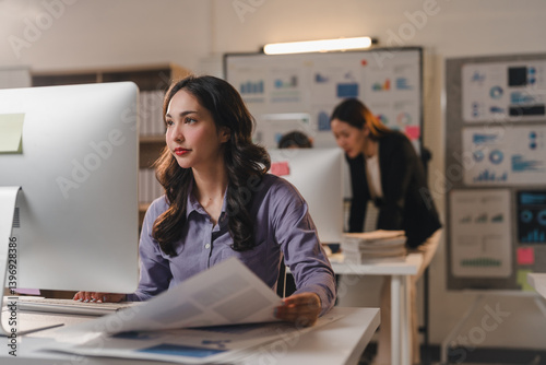 Young office worker analyzing marketing data on a computer while reviewing paperwork, with colleagues collaborating in the bustling background of a modern workplace