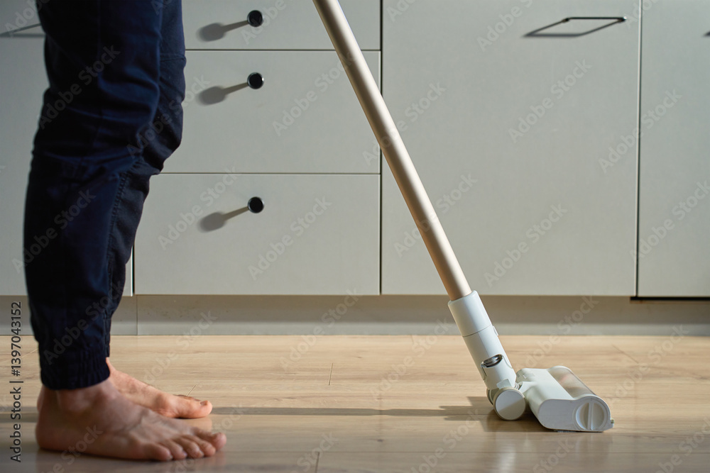 Barefoot man cleaning wooden floor in kitchen with vacuum. Person vacuuming with cordless vacuum cleaner. Daily routine and household chores