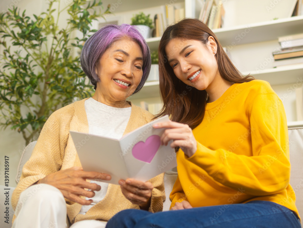 Cheerful Young asian woman daughter surprise her mother for mother's day holiday celebration giving postcard and gift while sits on sofa in living room at home. mother's day celebrating anniversary. Cheerful Young asian woman daughter surprise her mother for mother's day holiday celebration giving postcard and gift while sits on sofa in living room at home. mother's day celebrating anniversary.