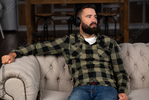 A bearded man sitting on a sofa, wearing headphones, lost in thought. Deep thinking, relaxation, personal reflection, or moments of contemplation.