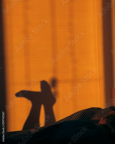 Casting shadows of legs on wall, glowing with warm orange light at sunset, creating cozy and relaxing atmosphere with person lying on bed, embracing tranquility. Sunset lit bedroom with outlined legs