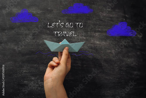 Hand holding a paper boat on a chalkboard with the words Let's go to travel