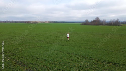 A young child joyfully explores a wide green field surrounded by distant trees. The sky is overcast, reflecting a serene autumn day. The scene captures a sense of adventure and freedom