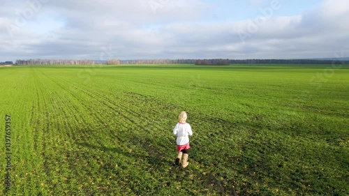 A young child joyfully explores a wide green field surrounded by distant trees. The sky is overcast, reflecting a serene autumn day. The scene captures a sense of adventure and freedom