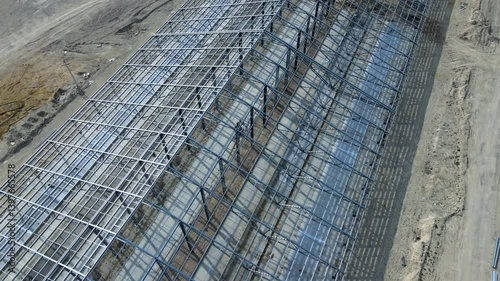 Flight over large metal building structure is taking shape in an agricultural zone. Metal frames are installed, creating an expansive space for future cultivation growth or animal husbandry