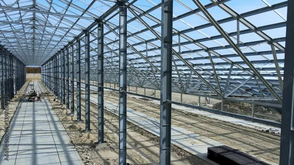 Flight inside the large metal building structure is taking shape in an agricultural zone. Metal frames are installed, creating an expansive space for future cultivation growth or animal husbandry Flight inside the large metal building structure is taking shape in an agricultural zone. Metal frames are installed, creating an expansive space for future cultivation growth or animal husbandry