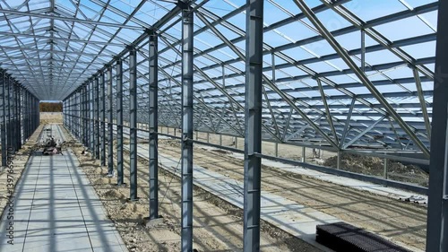 Flight inside the large metal building structure is taking shape in an agricultural zone. Metal frames are installed, creating an expansive space for future cultivation growth or animal husbandry