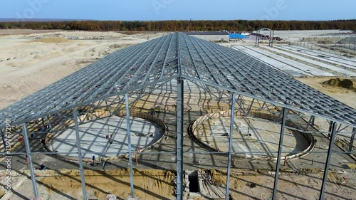 Flight over large metal building structure is taking shape in an agricultural zone. Metal frames are installed, creating an expansive space for future cultivation growth or animal husbandry