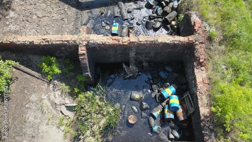 An abandoned industrial site shows waste and debris scattered amid overgrown vegetation. The polluted bitumen area highlights the impact of neglect on the environment