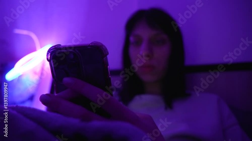 Woman holding a smartphone while lying in bed under purple neon lighting.