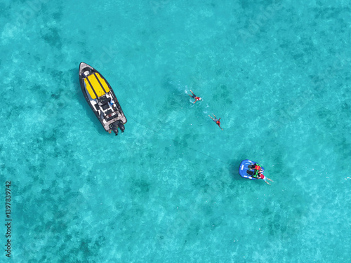 Aerial view of people enjoying water sports on a funtube behind a boat in turquoise clear water with beautiful corals, Vashafaru, Haa Alif, Maldives.