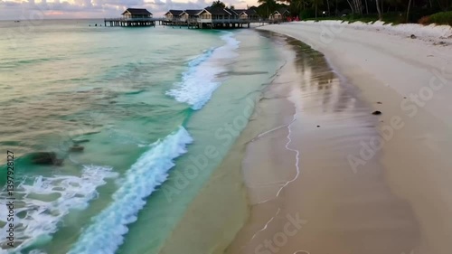 Crystalline turquoise waves gently caress the pristine sandy beach of a luxury overwater bungalow resort, creating a serene and idyllic tropical paradise.
