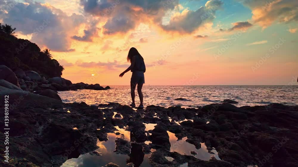 A girl stands on a rocky shoreline with her arms outstretched, embracing the beauty of a vibrant sunset over the ocean. The dramatic clouds and golden hues create a serene and uplifting atmosphere