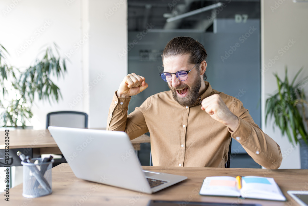 Fototapeta premium A man celebrates a success, beaming with joy as he looks at his laptop at his desk in the office, excited.