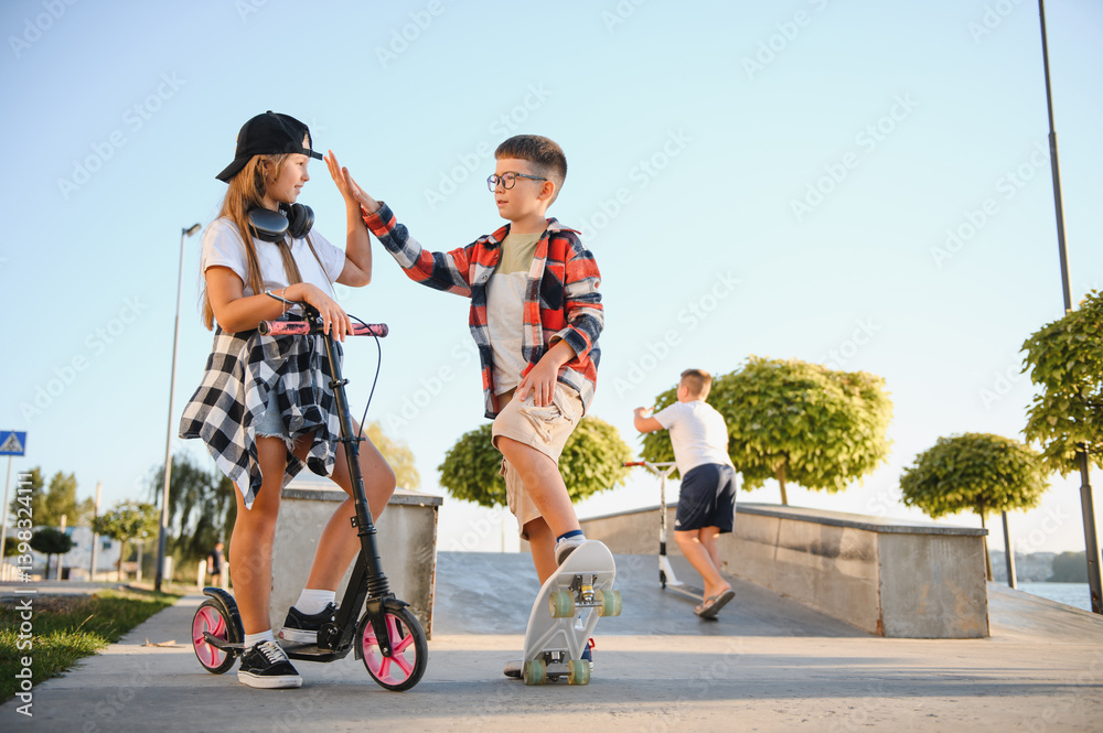 Fototapeta premium Cute kids in stylish casual clothes in skate park. They ride on skates and scooters. Summer vacation