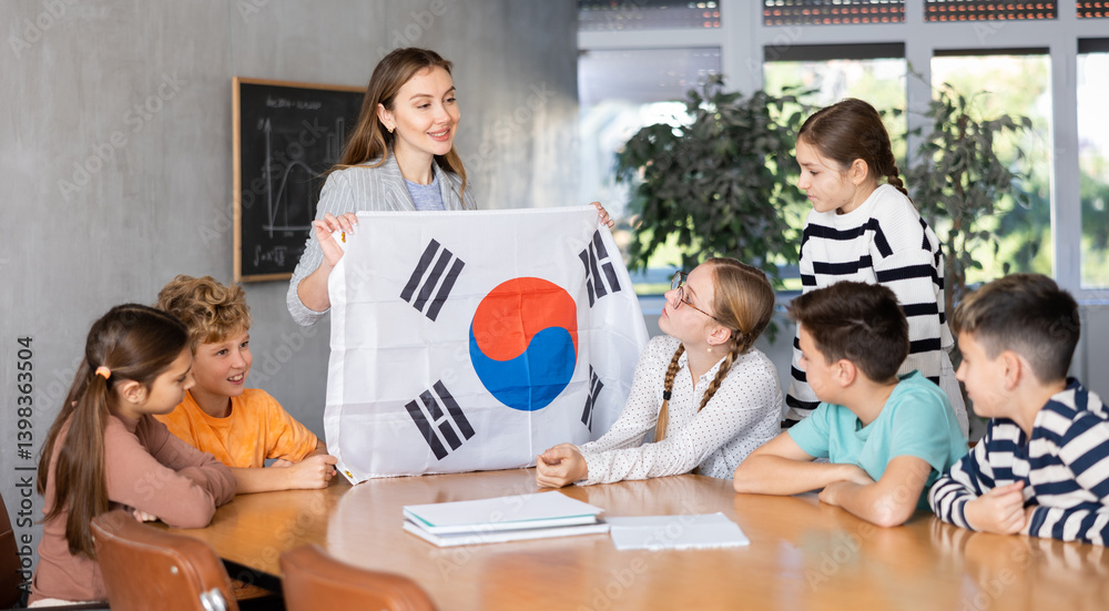 Joyful young female teacher showing flag of South Korea to schoolchildren preteens during history lesson in classroom