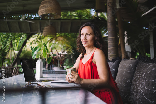 Obraz na płótnie Woman in red dress holding a coffee cup, looking thoughtful while seated at a laptop in an outdoor workspace with lush greenery. Relaxed, creative setting with natural decor and bohemian vibe w salonie
