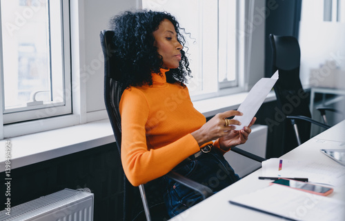 Confident and experienced businesswoman in orange sweater leans back in office chair, attentively reviewing document. Female focused expression reflects professionalism and expertise in her work