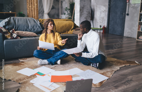 Obraz na płótnie Engaged couple sitting on the floor discussing documents, collaborating on finances or work projects. Surrounded by paperwork, they share a serious, focused moment in their cozy living room w salonie