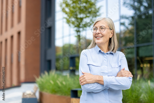 Bild auf Leinwand A smiling, mature businesswoman stands outdoors with arms crossed, near an office building