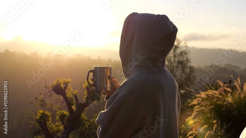 Back view of hooded woman drinking a hot beverage while enjoying a breathtaking sunrise over a mountain range, creating a serene and inspiring moment. Concept morning in mountains with cup of coffee