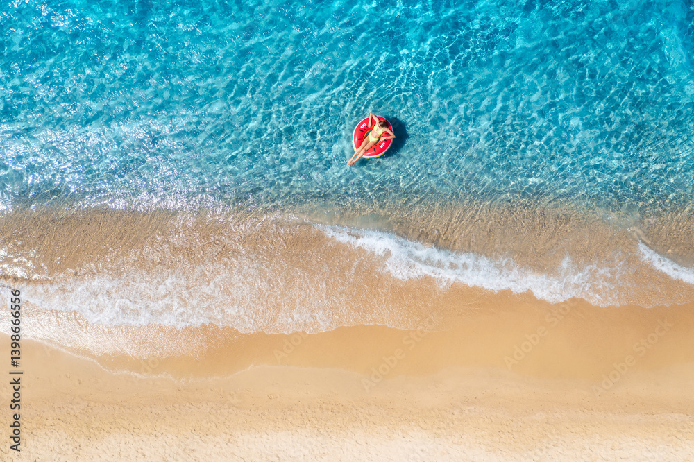 Naklejka premium Aerial view of a young woman swimming with red swim ring in blue sea with waves at sunset in summer. Tropical landscape with girl, clear water, sandy beach. Top view. Vacation. Sardinia island, Italy