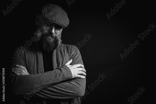 Obraz na płótnie Black and white, portrait of an emotional bearded man in a gray hat, gray sweater and glasses looking at the camera on a dark background. Dramatic portrait in dim lighting, expressive face, emotional w salonie