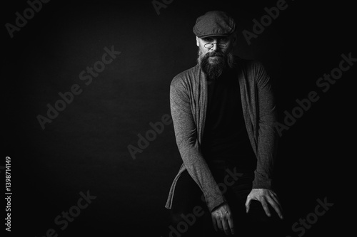 Obraz na płótnie Black and white, portrait of an emotional bearded man in a gray hat, gray sweater and glasses looking at the camera on a dark background. Dramatic portrait in dim lighting, expressive face, emotional w salonie