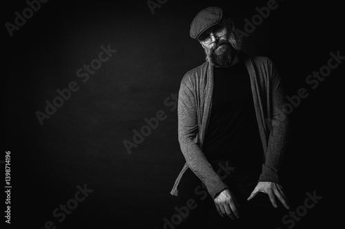 Obraz na płótnie Black and white, portrait of an emotional bearded man in a gray hat, gray sweater and glasses looking at the camera on a dark background. Dramatic portrait in dim lighting, expressive face, emotional w salonie
