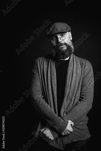Obraz na płótnie Black and white, portrait of an emotional bearded man in a gray hat, gray sweater and glasses looking at the camera on a dark background. Dramatic portrait in dim lighting, expressive face, emotional w salonie