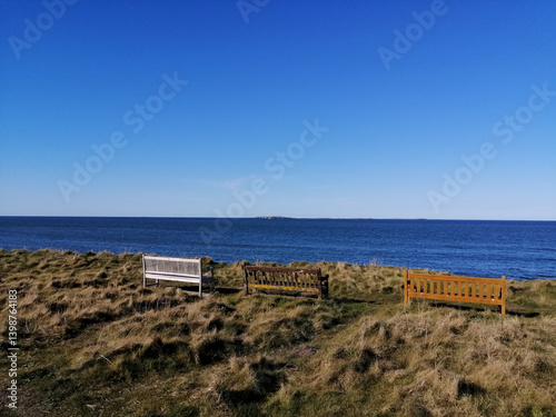 Three benches sit on a grassy hill, overlooking the beautiful blue sea on a sunny day.