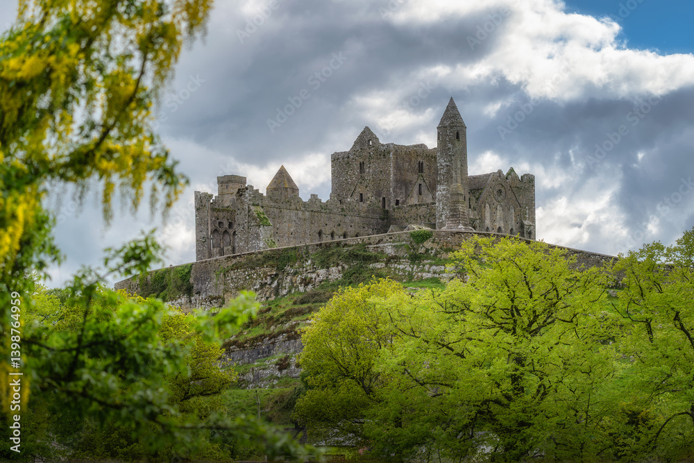 The breathtaking view showcases the Rock of Cashel medieval castle ruins majestically positioned on a prominent hill, beautifully framed by lush green trees beneath a dramatic sky, Tipperary, Ireland The breathtaking view showcases the Rock of Cashel medieval castle ruins majestically positioned on a prominent hill, beautifully framed by lush green trees beneath a dramatic sky, Tipperary, Ireland