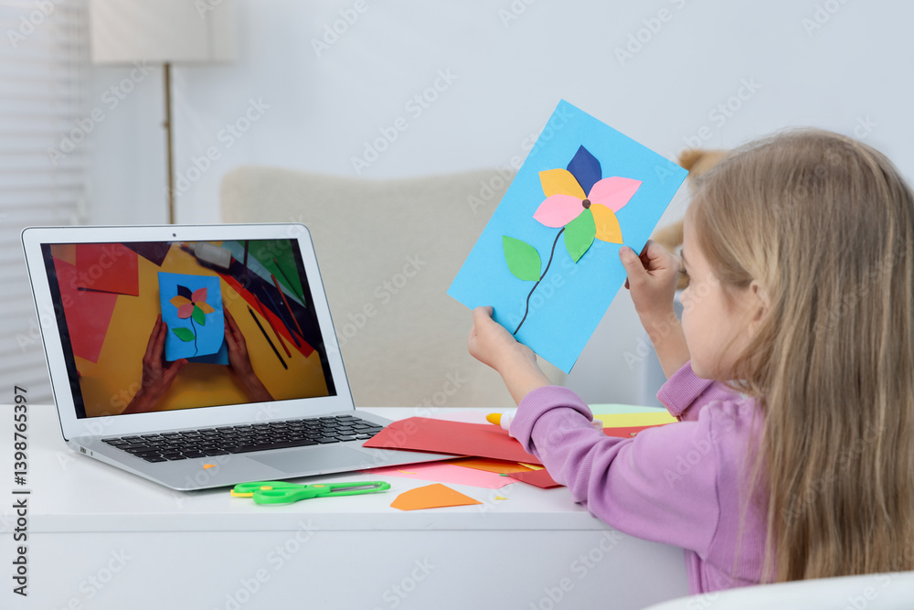 Little girl with handmade greeting card watching video tutorial at table indoors, back view
