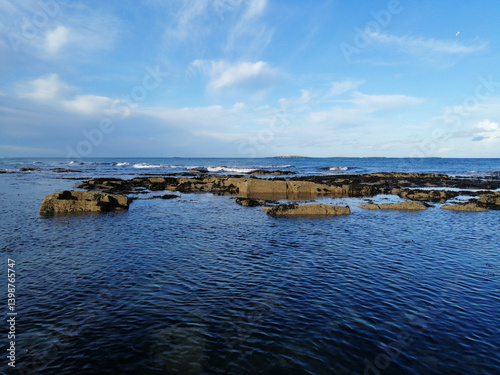A serene ocean view with rocks along the coastline under a bright blue sky with some clouds.