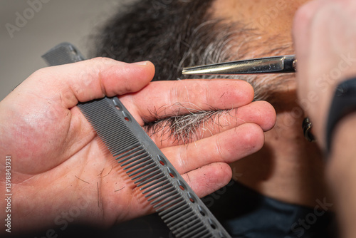 Close-up of a barber trimming a beard with electric clippers in a modern barbershop. Professional men's grooming and beard care service, selective focus	
