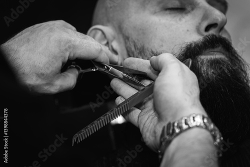 Black and white classic vintage photo, close-up, barber trimming beard with electric clipper, hair scissors in modern barbershop. Professional men's grooming and beard care services, selective focus