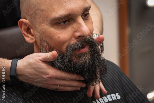 Obraz na płótnie Bearded man at barbershop getting beard groomed and shaped by barber. Close-up of grooming process, professional beard care, stylish male beauty routine w salonie