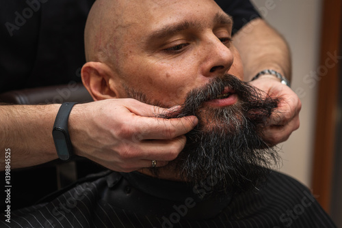 Obraz na płótnie Bearded man at barbershop getting beard groomed and shaped by barber. Close-up of grooming process, professional beard care, stylish male beauty routine w salonie