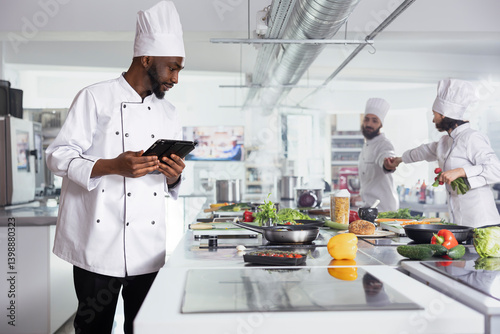 Billede på lærred Black head chef in a restaurant kitchen uses laptop to refine recipes, while kitchen tools and fresh ingredients fill the workspace for gourmet food prep