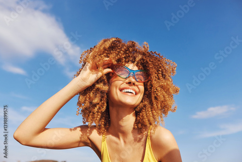 Smiling woman with curly hair wearing sunglasses against a bright blue sky
