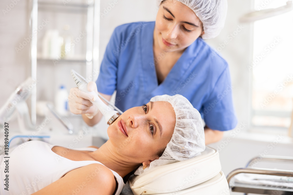 Obraz premium Young woman undergoing device-driven cosmetic facial treatment using high frequency ultrasound attachment stimulating opening of skin pores for deep penetration of moisturizers and nutrients