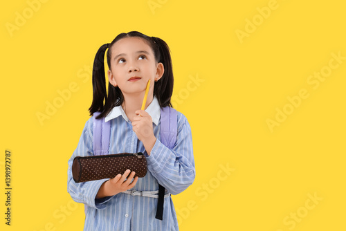 Cute little thoughtful Asian girl with backpack and pencil case on yellow bac...