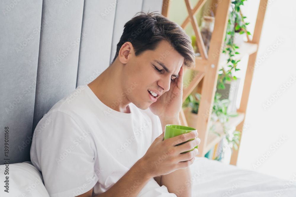 Young man in sleepwear holding a mug, sitting on bed at home, seeming contemplative during a morning in a cozy and bright bedroom Young man in sleepwear holding a mug, sitting on bed at home, seeming contemplative during a morning in a cozy and bright bedroom